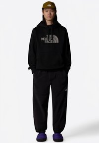 Sweatshirt preta com capuz e logótipo cinzento, calças pretas de corte largo e sapatos simples roxos, completado com um boné verde com logótipo.