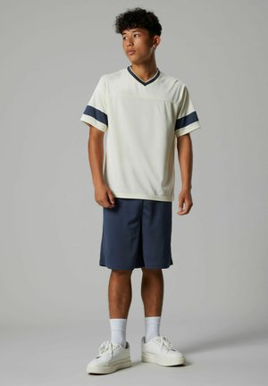 Young man standing wearing white V-neck shirt with dark stripes on sleeves, navy shorts, white socks, and white sneakers against gray background.