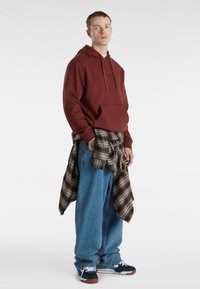 Felpa bordeaux con tasca frontale e coulisse, abbinata a jeans blu larghi e una camicia a quadri annodata. Sneakers blu scuro con dettagli bianchi.