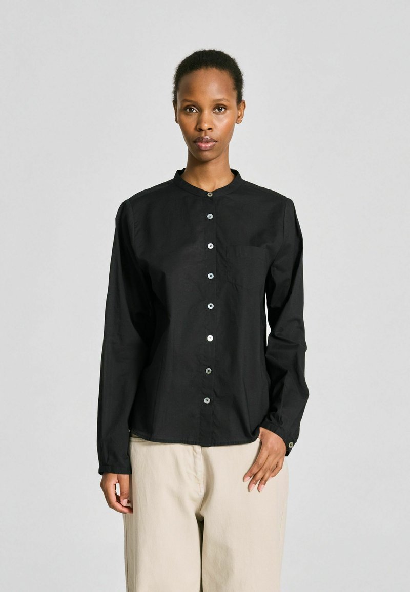 Personne debout face à l'avant, portant une chemise noire à boutons à manches longues et un pantalon beige à jambes larges, sur un fond clair uni.