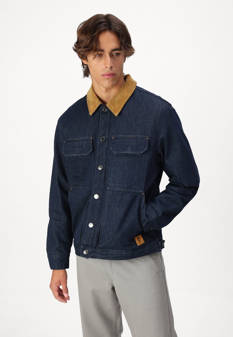 Jeansowa kurtka z musztardowym kołnierzem z korduroyu, z dwiema kieszeniami na piersi, zatrzaskami oraz brązową naszywką z logo po lewej stronie.