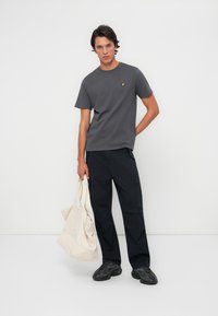 Szary t-shirt krótki rękaw z żółtym logo, w połączeniu z czarnymi spodniami. Model trzyma beżową torbę, nosząc czarne sneakersy o fakturze.