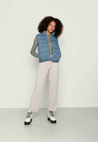 Colete acolchoado azul claro sobre um suéter cinza texturizado, combinado com calças bege claras e botas brancas com solas pretas.