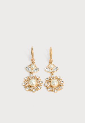 Božićne naušnice zlatne boje s centralnim biserima, prozirnim kristalnim laticama i malim kristalima koji tvore cvjetni uzorak na jednobojnoj svijetloj pozadini.