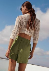 Gehaakte crème kleurige cardigan met lange mouwen, gecombineerd met olijfgroene linnen shorts met hoge taille en achterzakken. Helderblauwe lucht op de achtergrond.