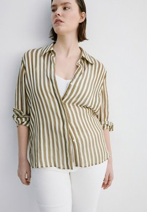 Mujer de cabello corto, vestida con pantalones blancos y una camisa de botones a rayas verticales en color oliva y blanco, con las mangas remangadas.