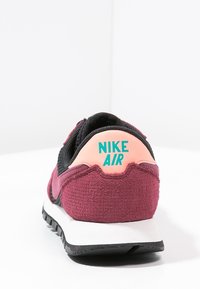 Bakifrån av en mörkröd och svart Nike Air sneaker med persiko-färgad hälflik och teal-färgad logotyp på en vit yta.