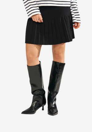 Donna con gonna nera plissettata, stivali neri lucidi alti fino al ginocchio e maglia a righe manica lunga, in piedi contro uno sfondo bianco.