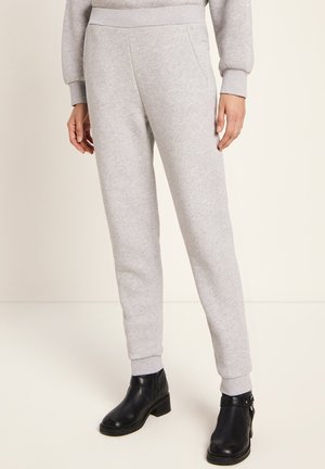 Person, die hellgraue, schmal geschnittene Jogginghosen mit seitlichen Taschen und schwarze Stiefeletten trägt, steht vor einem einfarbigen, hellen Hintergrund.