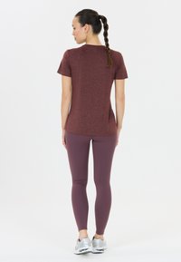 Vinröd sporttröja med korta ärmar, matchad med lila leggings. Modellen har rakt hår i fläta och står framför en enfärgad bakgrund.