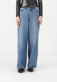 As calças de ganga azul de perna larga apresentam um tom claro, um design clássico de cinco bolsos e um corte descontraído, combinadas com uma blusa preta texturizada.