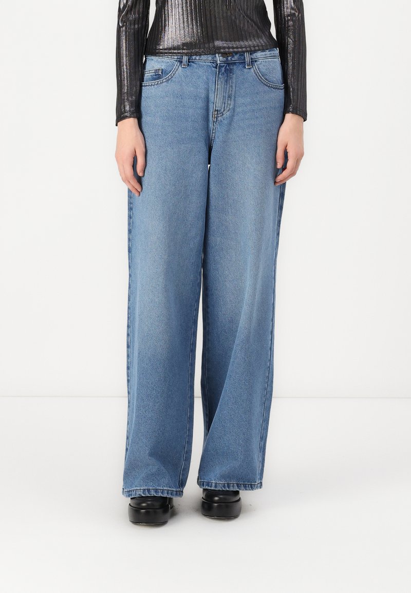 As calças de ganga azul de perna larga apresentam um tom claro, um design clássico de cinco bolsos e um corte descontraído, combinadas com uma blusa preta texturizada.