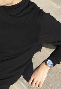 Camisa negra de manga larga, reloj de pulsera plateado con un dial azul y correa metálica, reposando sobre una superficie de piedra.