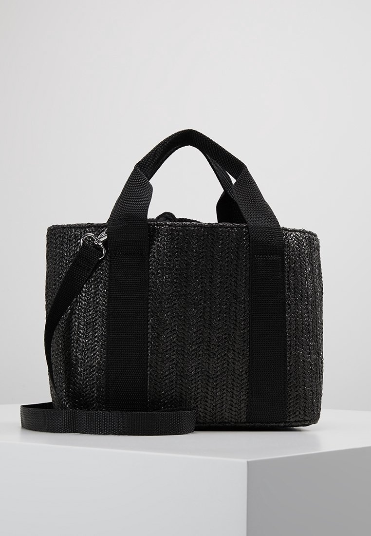 Bolso tote tejido negro con asas de tela negra gruesa y correa desmontable. Presenta una superficie texturizada con un patrón de espiga repetido.