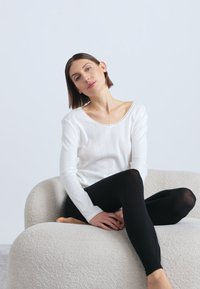 Λευκό ριμπ μακρυμάνικο τοπ με φαρδύ ντεκολτέ, συνδυασμένο με μαύρο κολάν, καθισμένη σε υφασμάτινο, ανοιχτόχρωμο καναπέ.