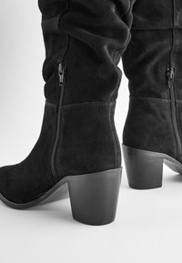 Bottes montantes en daim noir avec une coupe ajustée, fermetures éclair latérales et talons épais. La texture est douce, avec une finition lisse et un minimum de détails.