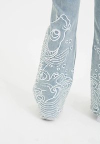 Himmelblå denim flared bukser med hvid broderede fisk og bølger langs de nederste ben, med en tekstureret overflade.