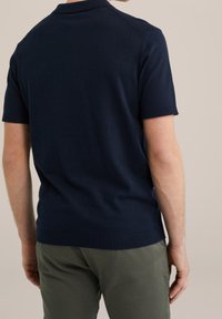 Námořnická pletená polo košile s krátkými rukávy, žebrovaným límcem a manžetami, hladkým povrchem a přiléhavým střihem, v kombinaci se světle zelenými kalhotami.