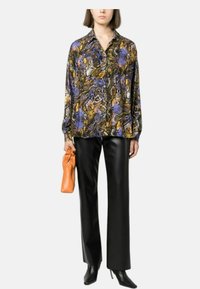 Blusa a maniche lunghe con motivo astratto multicolore in viola, oro e marrone; abbinata a pantaloni in ecopelle neri e borsa arancione.