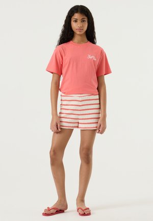 Koraalrood katoenen T-shirt met korte mouwen, gecombineerd met gestreepte crème- en oranje korte broek. Model draagt rode teenslippers. Eenvoudig, casual ontwerp.