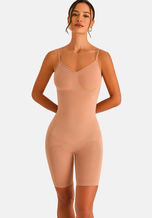Femme portant un body nude sans couture avec des fines bretelles s’étendant jusqu’au milieu de la cuisse, debout les mains derrière le dos sur fond blanc.