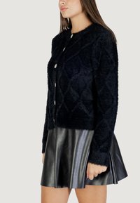 Cardigan noir en tricot duveteux avec un motif en losange, col rond et boutons en argent brillant. Porté avec une jupe évasée en cuir noir.
