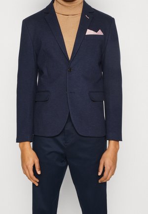 Mężczyzna w granatowej marynarce z różową poszetką, pod którą ma camelowy golf i granatowe spodnie, na jednolitym tle.