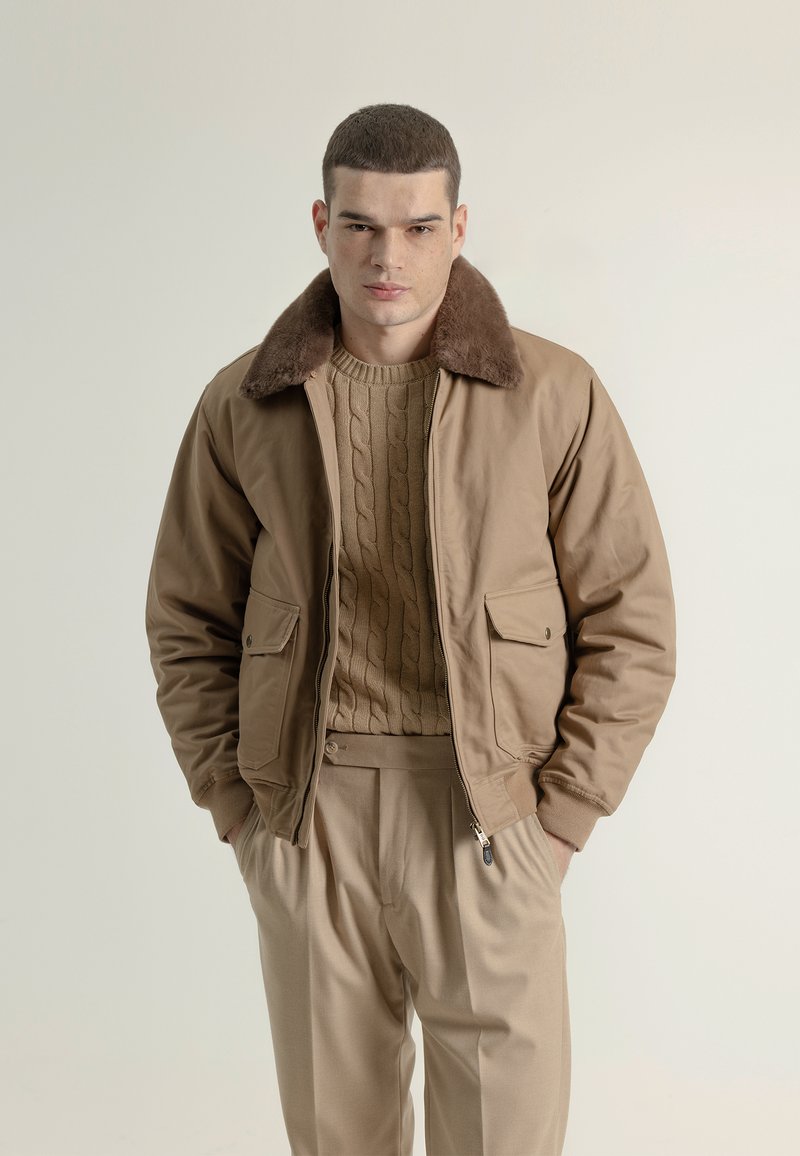 Chaqueta bomber marrón con cuello de piel, combinada con un suéter de punto beige y pantalones beige claro. Tejido suave con un diseño estructurado.