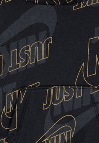 Jūras spēku kapucis ar atkārtotu zelta "TIKAI DARI TO" tekstu, ar priekšējo kabatu un mīkstu, teksturētu audumu. Melni akcenti fonā.