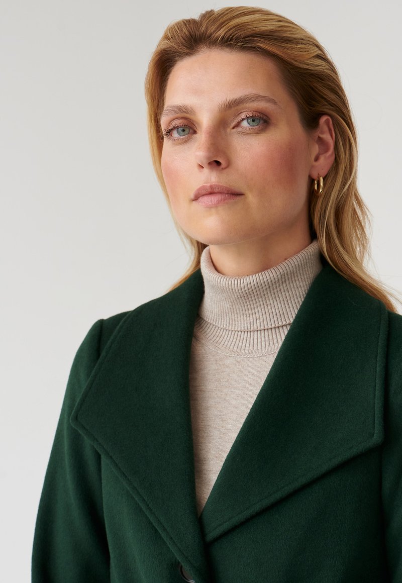 Cappotto di lana verde con ampi bavero, indossato sopra un dolcevita beige a coste. Presenta una texture liscia e una vestibilità su misura.