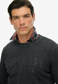 Hombre con cabello oscuro que lleva una sudadera negra de Superdry Athletics sobre una camisa de cuadros con cuello, mirando hacia adelante con una expresión neutral.