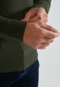 Pull en laine vert avec des poignets côtelés, présentant une texture douce et une coupe ajustée. Des mains ajustant le poignet sont visibles.