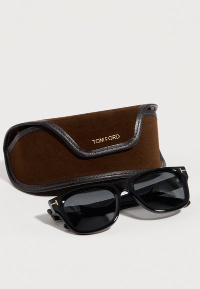 Μαύρα ορθογώνια γυαλιά ηλίου Tom Ford με χρυσές λεπτομέρειες λογότυπου δίπλα σε καφέ θήκη Tom Ford από σουέντ και δέρμα πάνω σε λευκή επιφάνεια.