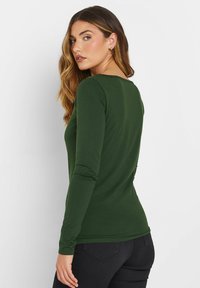 Grünes Langarmshirt aus weichem, dehnbarem Stoff mit rundem Halsausschnitt und figurbetontem Schnitt, kombiniert mit dunklen Jeans.
