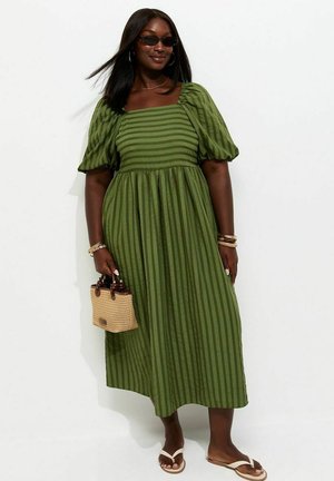 Vrouw in groene gestreepte midi-jurk met pofmouwen, zwarte zonnebril, houdt een rieten handtas vast, draagt armbanden en witte slippers.