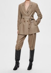Beige maatjasje met dubbele knopen, smalle taille en grote revers, in combinatie met losse beige broeken en kniehoge zwarte laarzen.