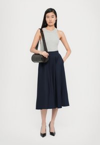 Graues ärmelloses Top, marineblauer plissierter Rock, schwarze spitze Pumps und eine runde schwarze Schultertasche. Glattes Material, hohe Taille und taillierte Passform.