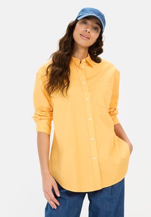 Geel overhem shirt met een borstzak, opgerolde mouwen en een gebogen zoom, gecombineerd met blauwe denim broek en een blauwe pet.