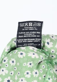 Grüner Stoff mit weißen Blumen und blauen Akzenten. Pflegeetikett angebracht, das Wasch- und Trocknungshinweise enthält. Weiche Textur, lässiges Design.
