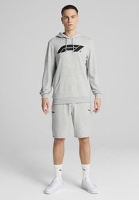 Hanorac gri cu glugă și logo negru, purtat cu pantaloni scurți gri, șosete albe și adidași albi. Un set athleisure casual cu o croială relaxată.