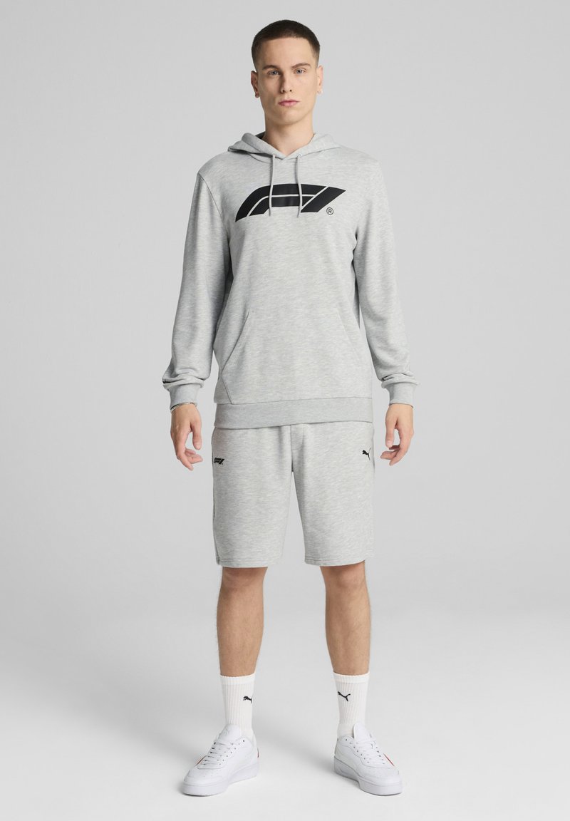 Hanorac gri cu glugă și logo negru, purtat cu pantaloni scurți gri, șosete albe și adidași albi. Un set athleisure casual cu o croială relaxată.