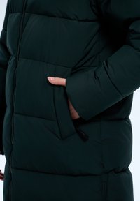 Puffet mørkegrønt jakke med lynlås, der har en sidelomme og en tekstureret overflade for isolering. Udsmykket med minimalistisk hardware.