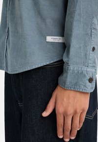 Lyseblå kordurhem med knap-ned krave, der har et lille mærketag, elastiske ærmer og en ribbebeskaffenhed. Mørk denim jeans synlige.