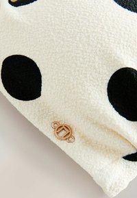 Material fleece alb cu buline negre și o etichetă mică rotundă din metal a mărcii atașată aproape de margine.