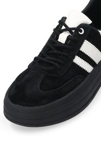 Czarne zamszowe sneakersy z białymi skórzanymi paskami, okrągłym noskiem i grubą czarną gumową podeszwą. Posiadają płaskie sznurowadła i wyściełany język.
