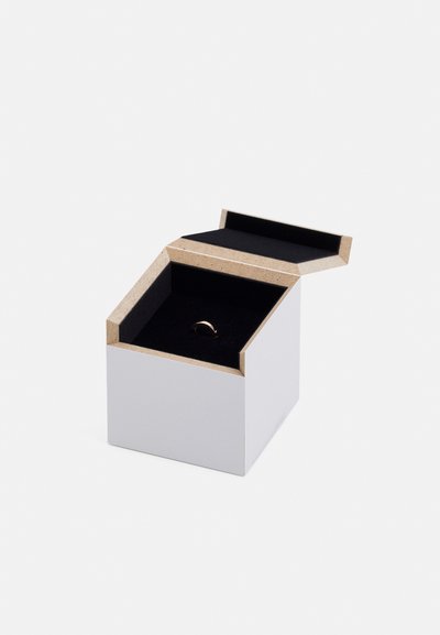 Cutie de bijuterii albă cu un capac unghiular unic, având un interior moale din catifea neagră și un inel de aur plasat în interior, evidențiind un design minimalist.