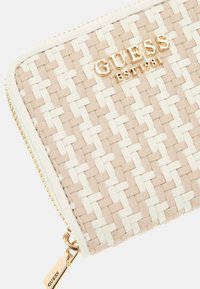 Woven beige and white wallet featuring a gold "GUESS" logo, gold zipper hardware, and a textured finish. Accented with a logo tag.