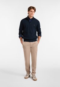 Suéter de punto navy con cuello con cremallera, combinado con pantalones beige claros y zapatillas beige. Presenta un pequeño logo en el pecho.