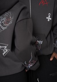 Grauer Kapuzenpullover mit grafischen Drucken, einschließlich detaillierter Designs an Ärmeln und Vorderseite. Verfügt über einen Reißverschluss und eine Kängurutasche.