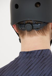 Svart hjälm med ventilationshål och justerbart harnesk, sedd bakifrån. Bäraren har kort hår och har på sig en mönstrad blå topp.